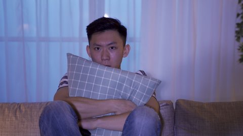 spooky ambience man holding pillow is frightened by the terrifying story. asian guy watching ghost movie let out a cry in fear when the evil spirit suddenly appears.