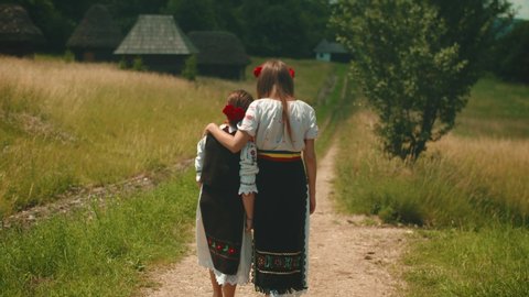 Two sisters dressed in traditional costume with red roses in their hair walking down a countryside road. Village life. Romania