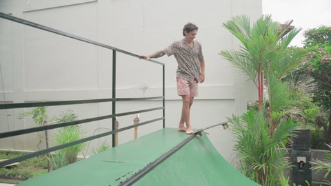 Handheld shot footage of active young man riding skateboard on half pipe in Bali villa yard loosing balance and falling down