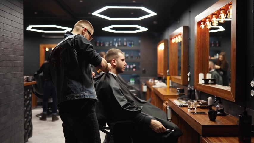 Trendy barber cuts bearded man's hair with a clipper in barbershop. Men's hairstyling and hair cutting in salon. Grooming the hair with trimmer. Hairdresser doing haircut in retro hair salon. Tracking | Shutterstock HD Video #1055208860