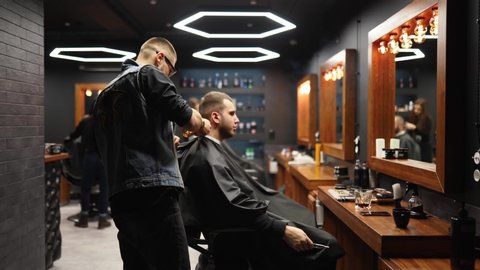 Trendy barber cuts bearded man's hair with a clipper in barbershop. Men's hairstyling and hair cutting in salon. Grooming the hair with trimmer. Hairdresser doing haircut in retro hair salon. Tracking