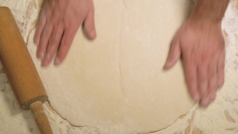 Male hands rolling dough. Top view, male hands roll out the dough with a rolling pin on a wooden surface, close-up. Close up of baker or chef preparing fresh dough with rolling pin on kitchen table.