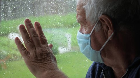 An elderly gray haired man in protective medical mask looks sadly out of window at the street during the rain, puts his hand on glass. Self isolation in a coronavirus pandemic, prevention of epidemic