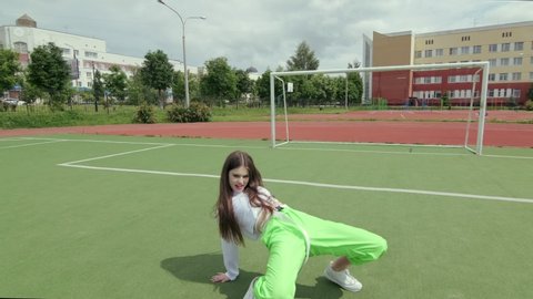 young woman with long hair in tracksuit dancing street modern dance, dancehall twerk, shaking her buttocks on a Sportsground. Urban dancing freestyle in the city. Booty Dance. Slow motion. Gimbal shot