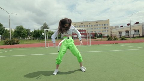 young woman with long hair in tracksuit dancing street modern dance, dancehall twerk, shaking her buttocks on a Sportsground. Urban dancing freestyle in the city. Booty Dance. Slow motion. Gimbal shot