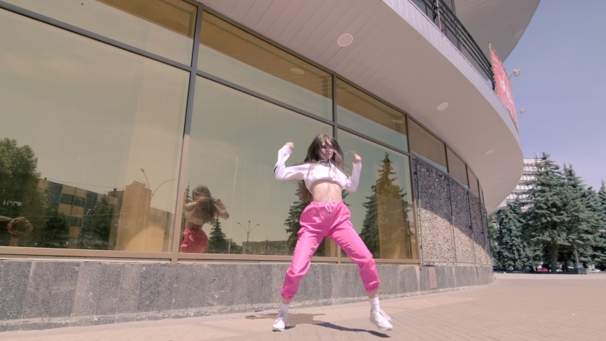 Funny girl performing cool dance opposite the building with mirrored glass. Concept of learning hip hop and break dance technique. Youth subculture. Contemporary choreography. Gimbal shot. Slow motion | Shutterstock HD Video #1055230499
