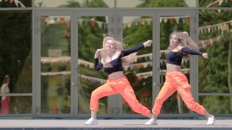 duet of two young girls with long hair in tracksuits dancing dancehall twerk, street modern dance in front of the building. Booty Dance. Urban dancing freestyle in the city. Slow motion