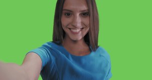 Young smiling woman in blue dress posing on green screen background. Girl taking selfie self portrait photo on smartphone. Chroma key. Female model showing positive face emotions. 4k raw video footage