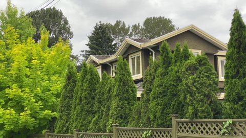 Establishing shot of two story stucco luxury house with green fence, big tree and nice landscape in Vancouver, Canada, North America. Dramatic clouds. Day time on June 2020. Pan right. H.264.