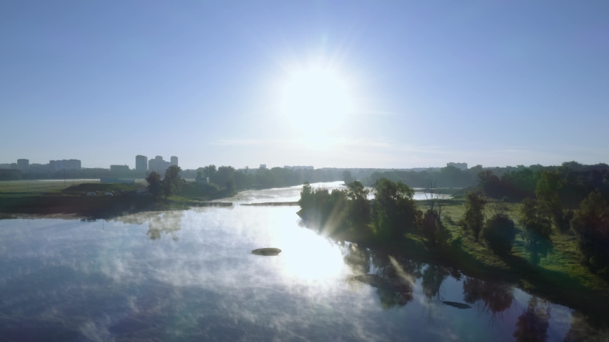 Aerial view of the river at dawn. The sky and the bright sun are reflected in the water. A white haze rises from the water.
