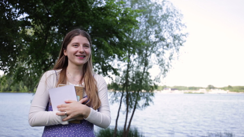 happy girl with books laughing and waving hand in park near river
