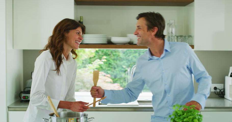 Authentic shot of a carefree happy smiling couple is listening to the music and having fun to dance and sing with a wooden spoon during cooking a dinner in the kitchen.  Royalty-Free Stock Footage #1055238311