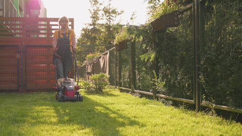Woman cutting grass with corded electric lawn mower in garden. Housewife mowing high green grass in private house yard. Outdoor seasonal household works.