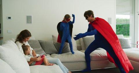 A carefree happy father and daughter dressed as superheroes are having fun to run and jump while playing an imagination game while mother with other daughter watching them and smiling at home.