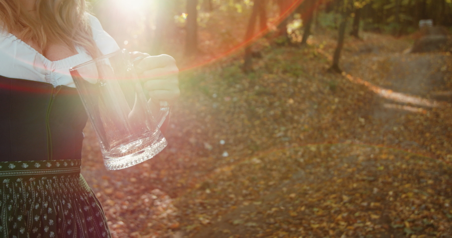 Slavic girl in national costume pours beer into a mug with sun beam in the the beautiful, autumn forest. Slowly 4K