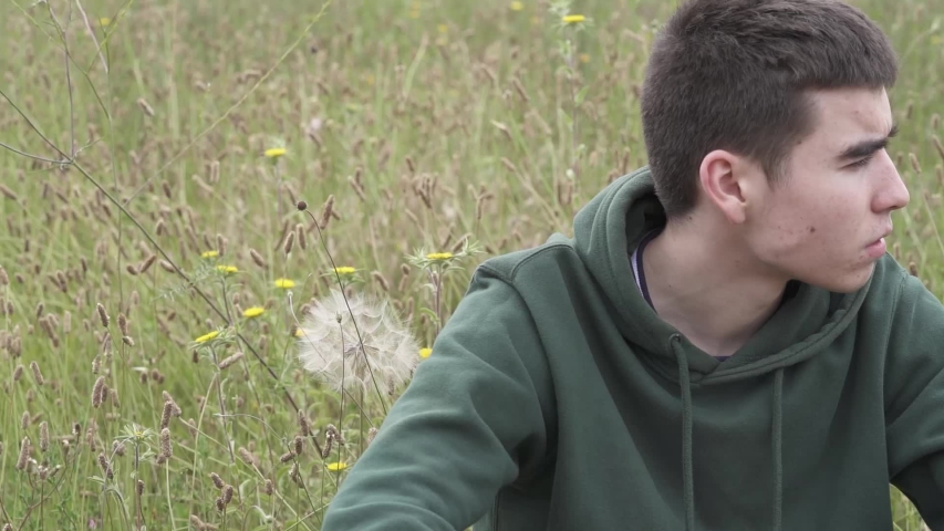 young guy sitting in a field and blowing out a dandelion
