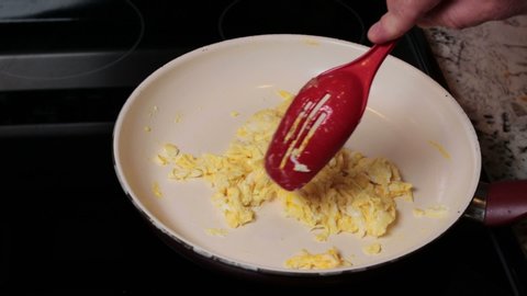 Organic free range scrambled eggs being cooked in a frying pan with a red spoon by a caucasian male hand. Finishing up stirring and cooking scrambled chicken egg yolks in a frying pan close-up.