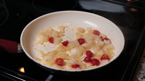 Male caucasian hand lifting non-stick pan and tossing white onions with grape tomatoes and placing bak on stovetop. Flipping white onions and grape tomato have in a frying pan cooking on the stovetop.