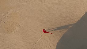 Nature and travel 4K aerial video. A young female in a beautiful fluttering red dress is walking by the desert landscape. Drone footage of a woman making footprints on the sand dunes at sunset, USA