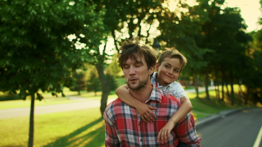 Closeup cheerful father giving son piggyback riding in park. Handsome man and boy talking on street. Adorable boy pointing with finger. Portrait of son and father looking up together