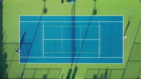 4K aerial view of the tennis serve. Drone shot of the match between two tennis players. Athletic family are practicing tennis at sunset. Game players are running and throwing the ball by rackets.