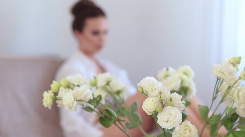 Blurred footage of young beautiful woman dressed in white lingerie and peignoir relax, lying on a bed in bedroom. Selective focus on a bouquet of white eustoma flowers, shallow depth of field.