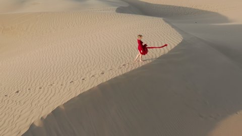Nature and travel 4K aerial video. A young female in a beautiful fluttering red dress is in the desert landscape. Drone footage of a woman making footprints on the sand dunes at sunset on a windy day : vidéo de stock