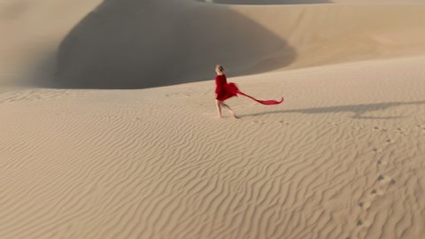 Nature and travel 4K aerial video. Lonely young woman in a beautiful fluttering red dress is walking by the desert landscape at sunset. Drone footage of a girl on a windy day at the sand dunes, USA