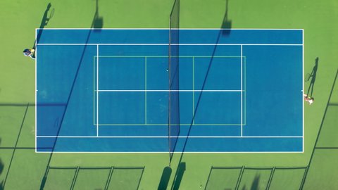 4K aerial view of the tennis match. Athletic father and daughter are practicing tennis at sunset time. Active game players are running by the court and throwing the ball by their rackets.