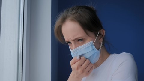 Slow motion: woman wearing face mask, feeling sick, suffering from coughing, looking out of window in room with blue wall. Self isolation, prevention, quarantine, COVID 19, coronavirus, safety concept