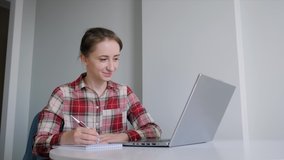 Slow motion: woman in red plaid shirt using laptop, having video call, making notes, talking in grey room. Education, e-learning, conference, self isolation, quarantine, online technology concept