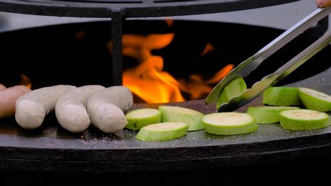 Slow motion: chef grilling fresh meat sausages and zucchini slices on black brazier with hot flame at summer food market - close up. Outdoor cooking, barbecue, gastronomy, cookery, street food concept