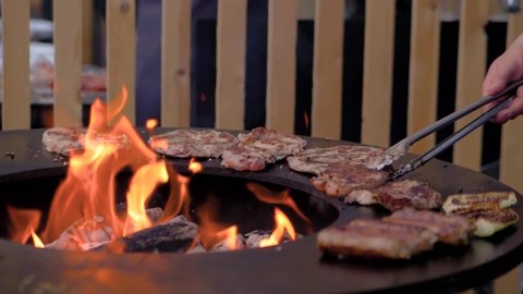 Super slow motion: chef grilling fresh meat steaks on brazier with hot flame at summer local food market - close up view. Outdoor cooking, barbecue, gastronomy, cookery, street food concept