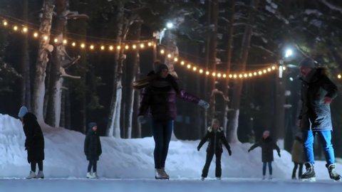 boyfriend lifts up girl in short jacket and skates having fun on skating rink with garlands at winter night slow motion