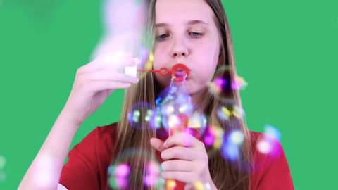 Close-up portrait of an attractive cheerful girl with long hair in red t-shirt blows some soap bubbles at the camera, smiling kindly, enjoying the moment. Isolated on green background.