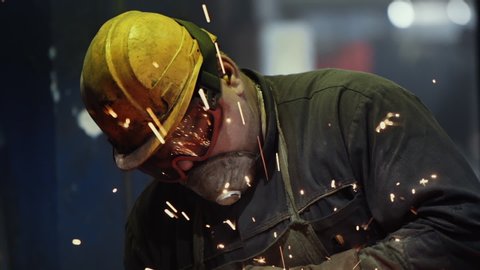 Tough Work of a Welder in Green Uniform and Protection Yellow Helmet at Steel Making Factory. Angle grinder at Metal Production. Sparks, Slow Motion, Close Up