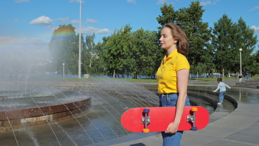 A teenage girl with a skateboard in her hands poses at the camera next to a fountain in a city park. The concept of urban life and youth culture
