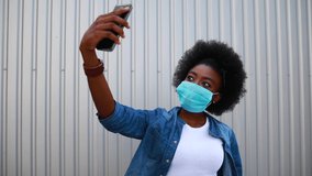Portrait of a pretty young african american student woman in medical mask. Makes a selfie on the phone. Black afro american girl in shirt with afro hairstyle. Portrait