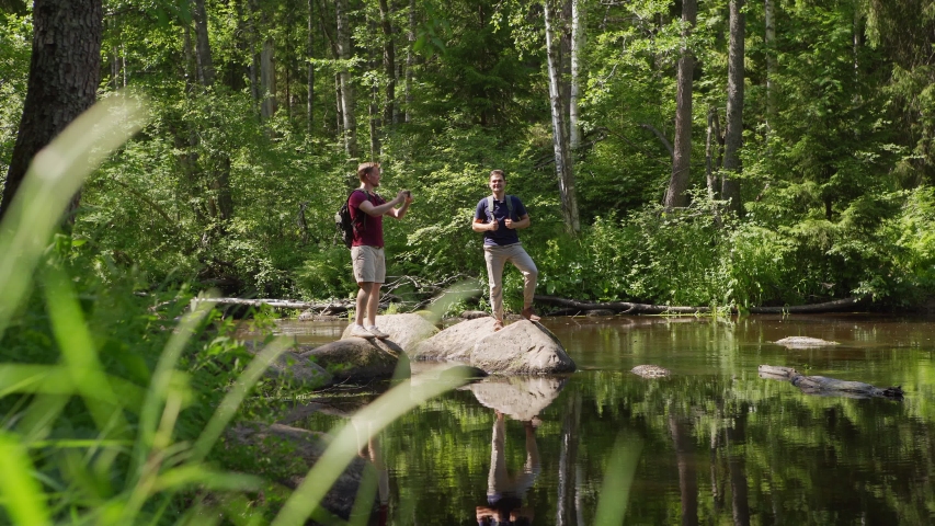 Tracking wide shot of two male hikers with backpacks jumping on rocks to photograph bautiful view over forest river on smartphone