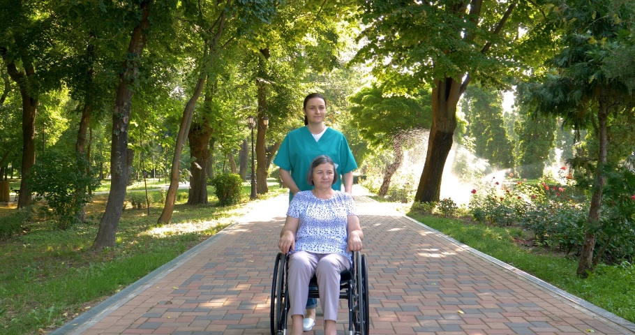 Young nurse spending sunny day with senior woman in wheelchair walking in park. 4k | Shutterstock HD Video #1055278985