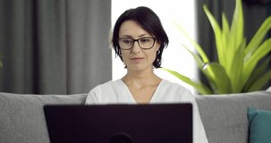 Positive mature woman with dark hair sitting comfortably on grey couch and using laptop. Casual female in eyeglasses staying at home and working on computer.