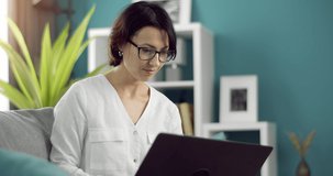 Confident dark-haired woman using portable laptop while sitting on couch and working. Attractive lady in eyeglasses spending time at home for remote work.
