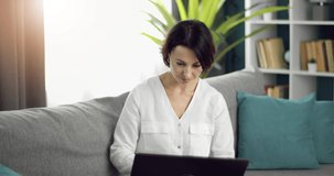 Charming lady with trendy haircut sitting on grey couch, smiling and looking at computer screen. Pretty woman in white shirt using wireless laptop at home.