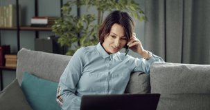 Woman with pleasant appearance talking on smartphone and using portable computer while sitting on grey sofa. Charming female in casual outfit staying at home with modern gadgets.