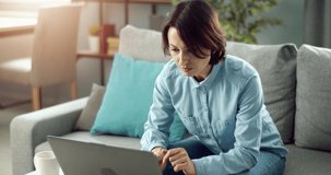 Concentrated woman with short haircut sitting on grey couch and using laptop. Female freelancer in denim shirt and jeans working remotely at home.