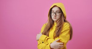 Cute young girl dressed in yellow raincoat with hood, warming herself during rainy day. Beautiful woman feeling cold because of bad weather. Isolated over pink background.