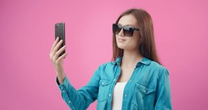Happy young girl with dark hair using smartphone for taking selfie. Stunning woman wearing trendy sunglasses and denim shirt. Isolated over pink studio background.