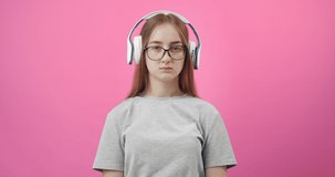 Sad young girl in eyeglasses and wireless headphones listening lyric song and suffering from loneliness. Isolated over pink studio background.