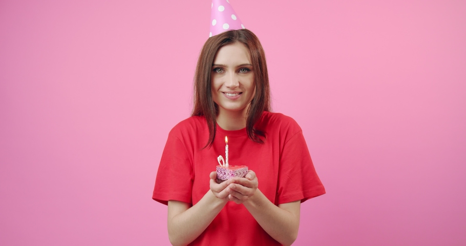 Happy beautiful girl wearing red t-shirt and birthday cap blowing burning candle on small cake that holding in hands. Young woman with dark hair making wish and enjoying birthday day.