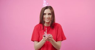 Happy beautiful girl wearing red t-shirt and birthday cap blowing burning candle on small cake that holding in hands. Young woman with dark hair making wish and enjoying birthday day.